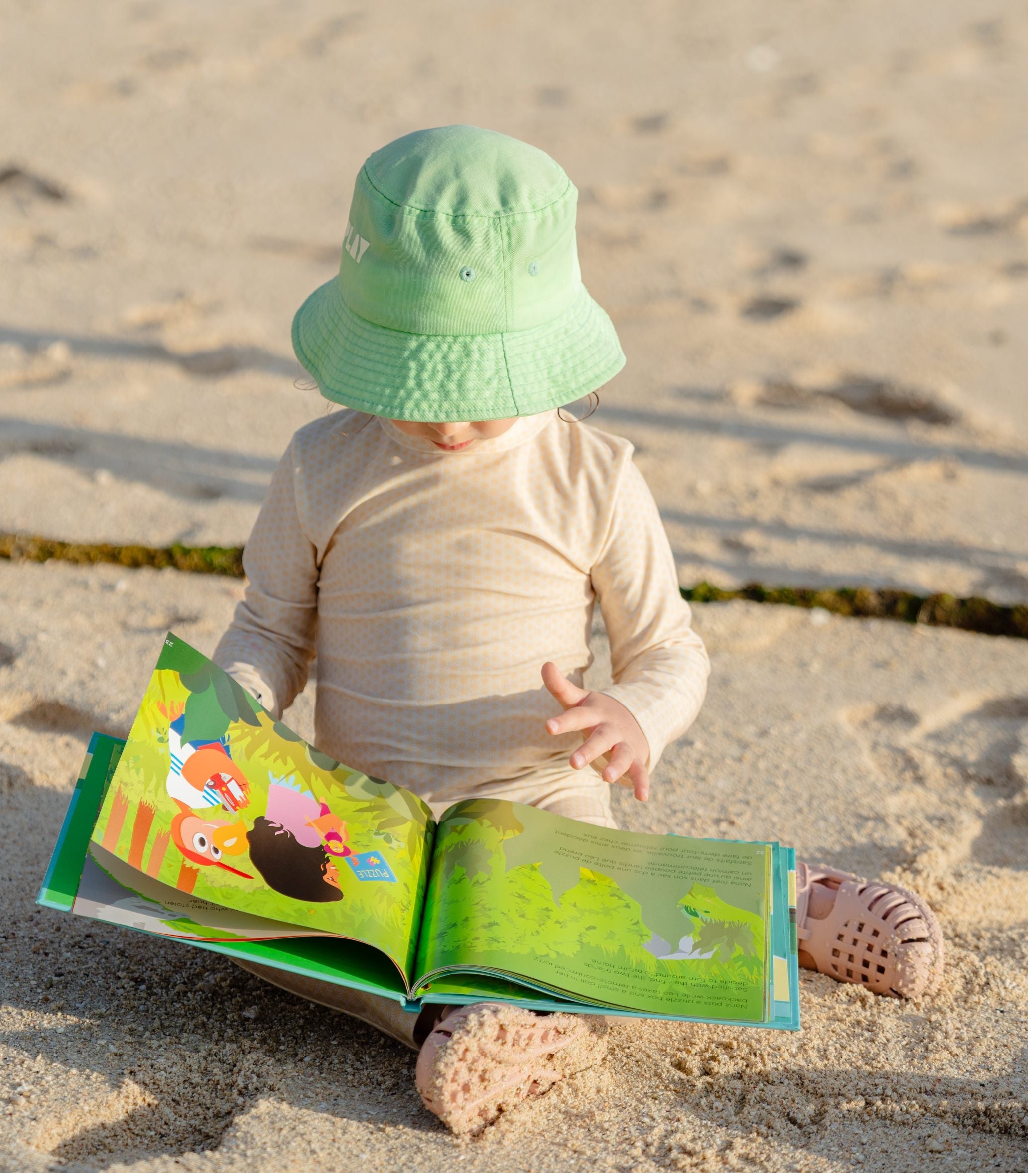 Toddler sitting on a beach, wearing a full coverage MIKOU sun suit, is looking at a colourful book about Dodo. 