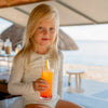 A young girl sitting on a beach bar counter, wearing a MIKOU UPF50+ UV Protective long sleeve swimsuit and drinking a colorful beverage.